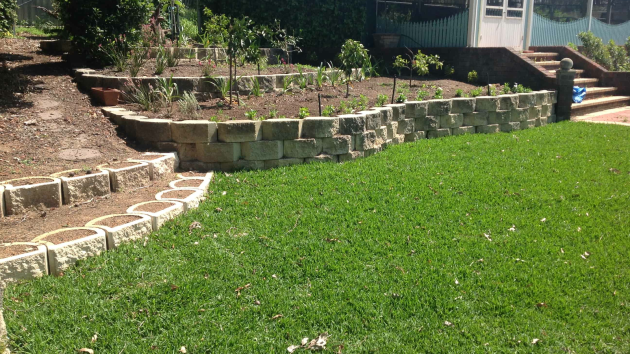 Tiered retaining walls built from textured blocks, with a curved block edge, young plants and a lawn in front